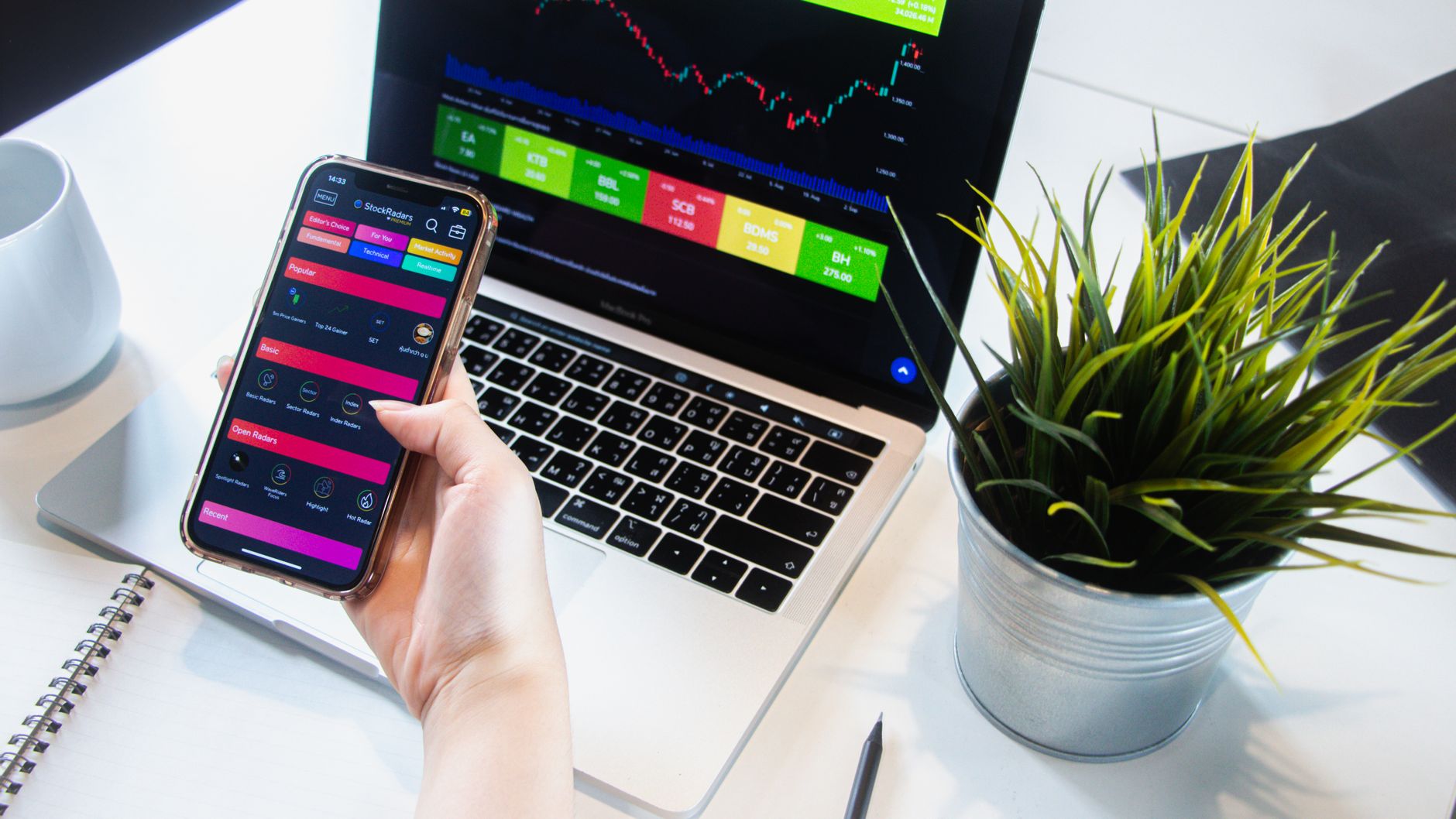 Ilustrasi: A person using a smartphone app to trade stocks with a laptop displaying market data.